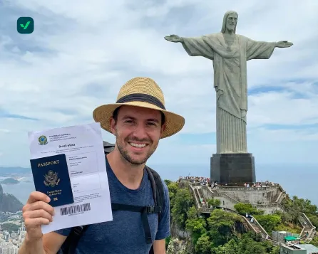 Smiling traveler wearing a hat holds a U.S. passport and a printed Brazil eVisa document, standing in front of the Christ the Redeemer statue in Rio de Janeiro.